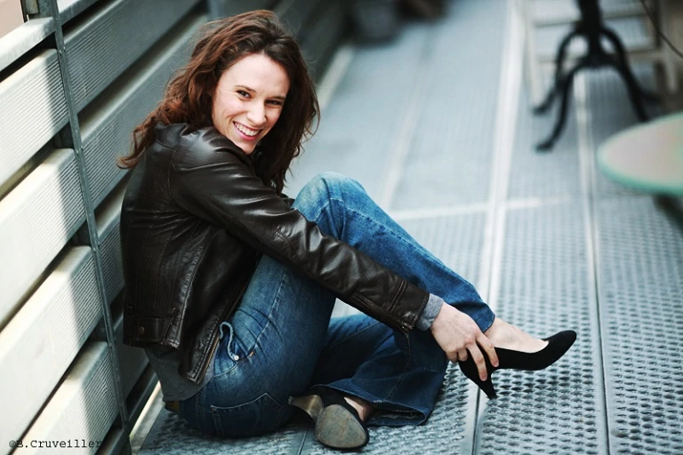 book photo une jeune femme brune cheveux détachés en jean et veste en cuir assise sur une plate forme en alu remet sa chaussure en regardant l'objectif elle sourit