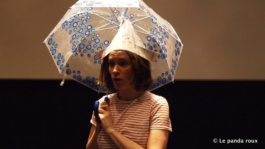 photo de spectacle une jeune comédienne brune les cheveux détachés avec une chapeau en papier journal s'abrite sous un parapluie elle a l'air triste