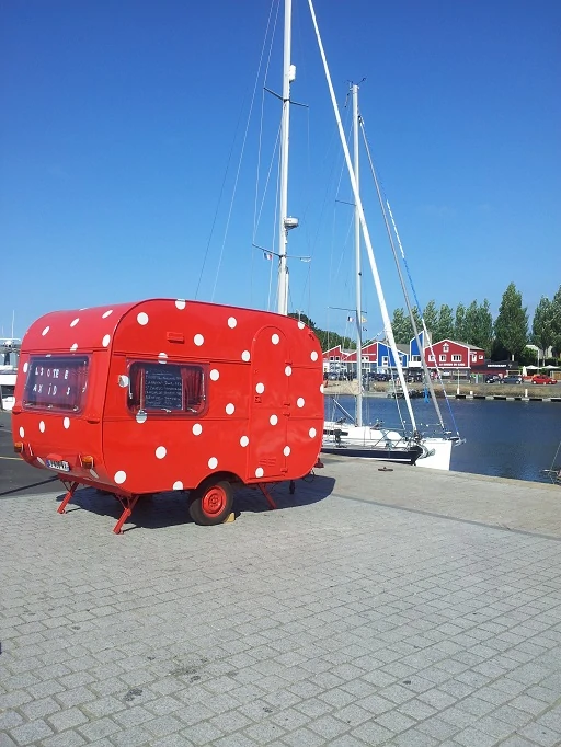 une caravane rouge à pois blancs est posée sur le port d'une ville avec les bateaux derrière il fait beau