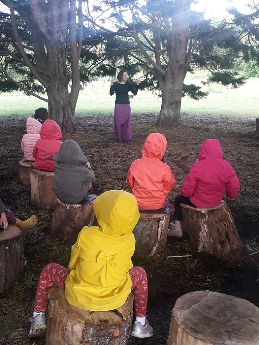 une conteuse debout raconte des histoires à des enfants assis sur des rondins et encapuchonnés sous la pluie dans leurs cirés colorés au milieu d'une clairière d'arbres