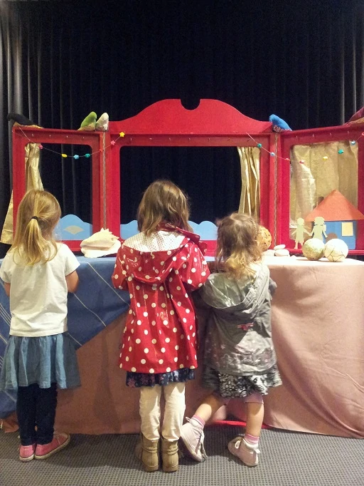 trois petites filles nous tournant le dos se sont approchées du castelet d'une comédienne après le spectacle et regardent les marionnettes