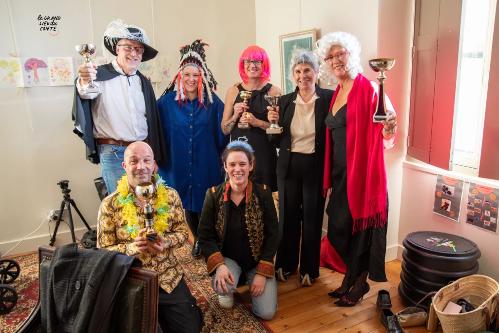 Un groupe de personnes pose pour la photo. Ils et elles sourient et portent des costumes loufoques mais élégants. Ils et elles tiennent des coupes en or et en argent dans les mains.