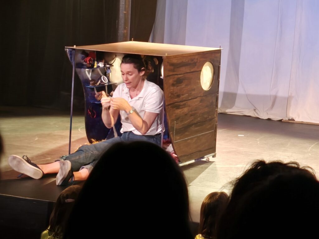 une comédienne est assise dans une petite boîte sur scène pendant un spectacle. On dirait un enfant. Elle joue avec des petits objets.