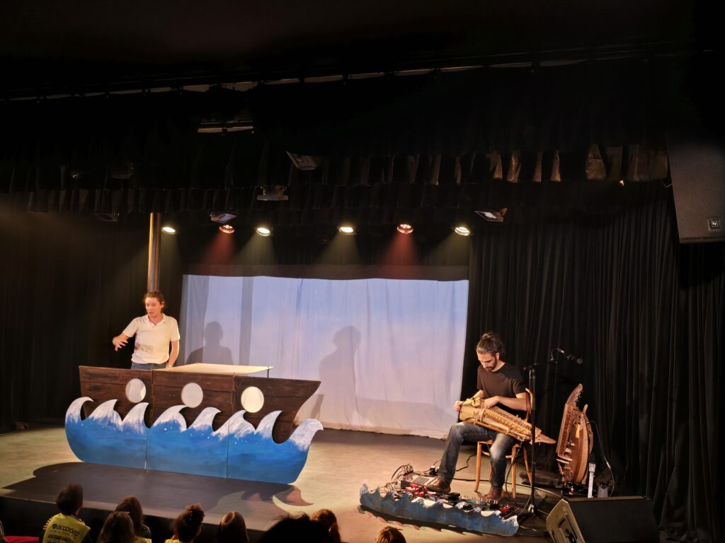 Pendant un spectacle sur scène, une comédienne se tient debout dans un bateau avec les vagues dessous. A côté d'elle, un musicien joue de,la vielle à roue, assis sur une chaise.