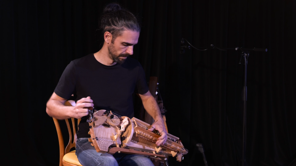 un musicien joue de la vielle à roue, assis sur une chaise. il a une quarantaine d'années et porte un tee-shirt noir et les cheveux longs.